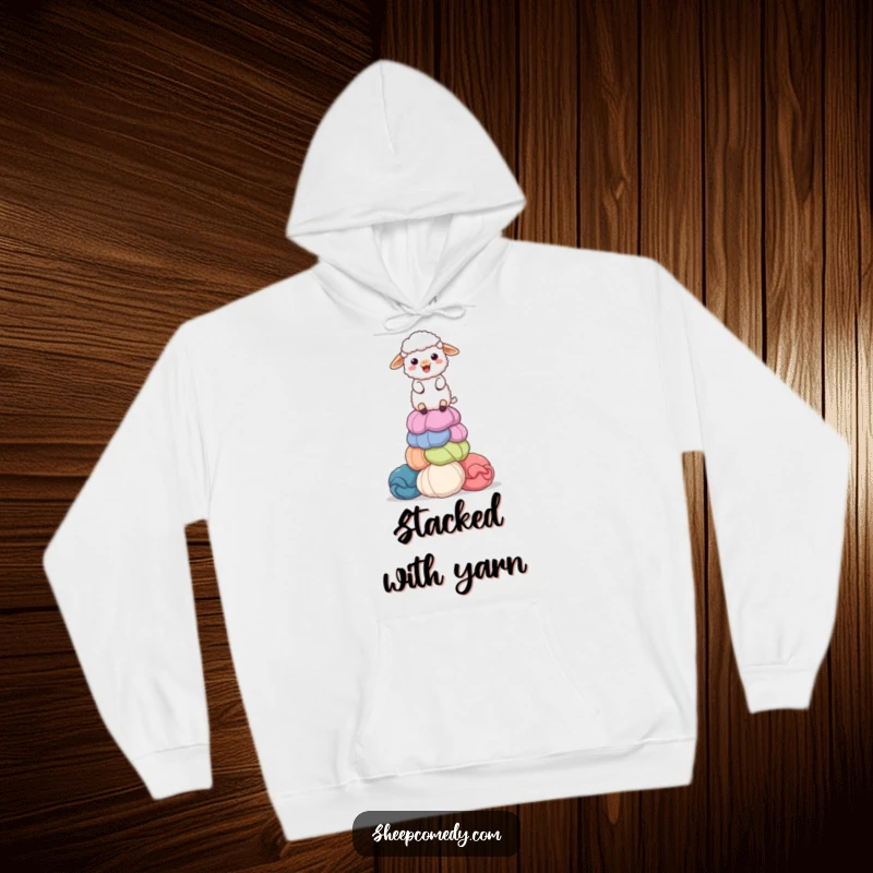 Funny hoodie depicting a delighted sheep balancing a stack of colorful, giant wool balls, ideal for cozy, humorous wear during crafting.
